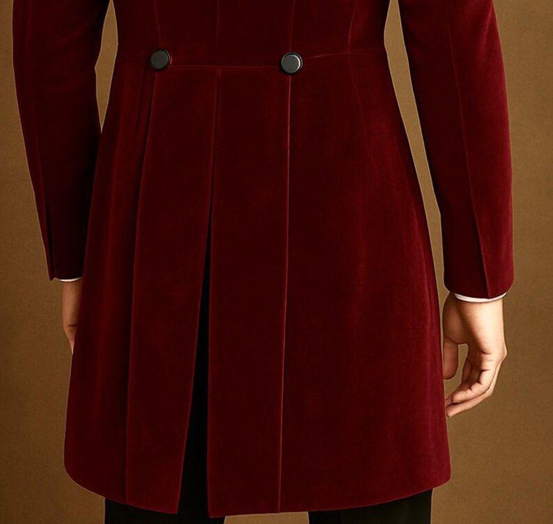 May include: A burgundy velvet coat with black buttons and a pleated back. The coat has long sleeves with white cuffs. The garment is a formal style.