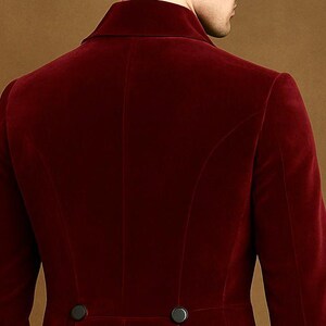 May include: A close-up of the back of a burgundy velvet blazer. The blazer has a notched lapel, two black buttons, and a tailored fit. The fabric has a soft, luxurious appearance. The background is a solid, warm brown.