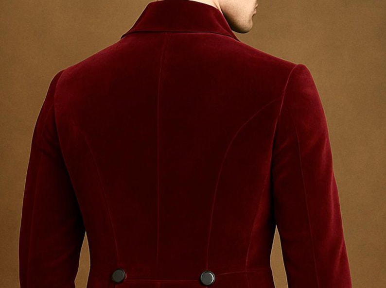 May include: A close-up of the back of a burgundy velvet blazer. The blazer has a notched lapel, two black buttons, and a tailored fit. The fabric has a soft, luxurious appearance. The background is a solid, warm brown.