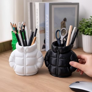 May include: Two puffer jacket-shaped pencil holders, one white and one black, each featuring a central zipper detail and a high collar. These containers are filled with various pens, pencils, and scissors, serving as functional desk organizers.