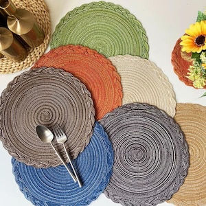 Op de afbeelding: Een verzameling kleurrijke ronde placemats in groen, oranje, bruin, beige, blauw en grijs. Elke placemat heeft een gevlochten rand en een spiraalvormig ontwerp. Een lepel en vork liggen op een van de placemats. De placemats zijn gemaakt van geweven materiaal.
