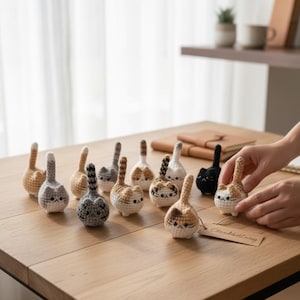 May include: A collection of crocheted cat figurines in various colors, including gray, brown, and black, with white accents. Each cat has a distinctive tail and facial features. The figurines are arranged on a wooden surface.