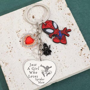 Puede incluir: Un llavero plateado con un dije de Spider-Man rojo y negro, un dije en forma de coraz&oacute;n rojo con un borde de diamantes de imitaci&oacute;n y un dije de ara&ntilde;a negro. Tambi&eacute;n se incluye un colgante en forma de coraz&oacute;n con el texto "Just A Girl Who Loves Spider Man".