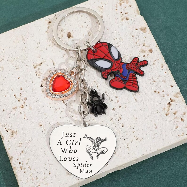 Puede incluir: Un llavero plateado con un dije de Spider-Man rojo y negro, un dije en forma de coraz&oacute;n rojo con un borde de diamantes de imitaci&oacute;n y un dije de ara&ntilde;a negro. Tambi&eacute;n se incluye un colgante en forma de coraz&oacute;n con el texto "Just A Girl Who Loves Spider Man".