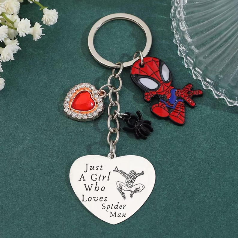 Puede incluir: Un llavero plateado con m&uacute;ltiples dijes. Los dijes incluyen una figura de Spider-Man roja y negra, una ara&ntilde;a negra, un dije en forma de coraz&oacute;n con un centro rojo y un colgante en forma de coraz&oacute;n con el texto "Just A Girl Who Loves Spider Man."