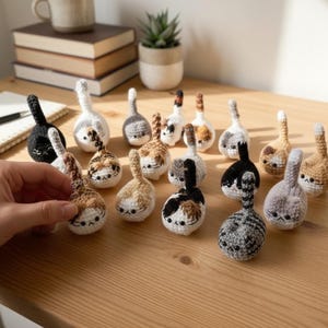 Op de afbeelding: Een verzameling gehaakte kattenkontornamenten in diverse kleuren en patronen, waaronder zwart, grijs, lapjeskat en tabby. Elk ornament heeft een gedetailleerd kattengezicht en staart.