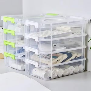 Op de afbeelding: Transparante, stapelbare opbergcontainers van plastic met witte en groene accenten. Elke container heeft meerdere planken gevuld met kantoorbenodigdheden, waaronder een rekenmachine, notitieboekjes en rollen tape. De containers zijn ontworpen voor het organiseren en opslaan van diverse items.