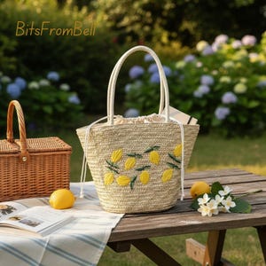 May include: A straw tote bag with embroidered yellow lemons and green leaves. The bag has white handles and a drawstring closure. A picnic basket, lemons, and flowers are on a wooden table. The text "BitsFromBell" is in the upper left corner.