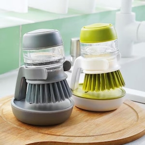 Könnte beinhalten: Zwei Geschirrbürsten mit integrierten Seifenspendern. Eine Bürste ist grau mit einem grauen Griff und Borsten, die andere ist weiß und grün mit grünen Borsten. Beide haben transparente Seifenbehälter und stehen auf einer runden Basis.