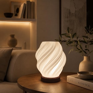 Può includere: Una lampada da tavolo in vetro bianco a spirale con una base in legno marrone scuro. La lampada è accesa, emettendo un bagliore caldo. Si trova su un tavolo rotondo di legno, accanto a un libro e un vaso con vegetazione. Il design della lampada presenta un motivo a spirale.
