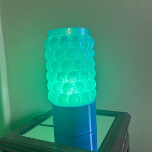 Può includere: Una lampada cilindrica con un design strutturato a forma di bolle, che emette un bagliore verde vibrante. La lampada è posizionata su una base blu e su una superficie di legno. Il design della lampada ricorda una serie di sfere impilate.
