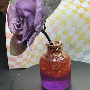 Puede incluir: Un pequeño jarrón decorativo con un degradado de colores, que va del naranja al morado. El jarrón está lleno de purpurina y contiene una rosa morada con hojas grises. El fondo presenta un patrón de cuadros.