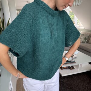 May include: A teal green, short-sleeved knit sweater. The sweater has a crew neck and a slightly oversized fit. The knit fabric appears to be a chunky yarn, giving it a textured appearance. The sleeves are short, ending just above the elbow.
