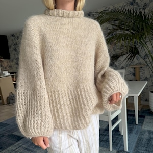 May include: A beige, oversized sweater with a ribbed turtleneck and balloon sleeves. The sweater has a textured knit design and a ribbed hem. The sleeves are gathered at the wrist. The sweater is made of a soft, warm material.