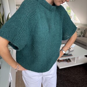 May include: A teal green, short-sleeved knit sweater. The sweater has a crew neck and a slightly oversized fit. The knit fabric appears to be a chunky yarn, giving it a textured appearance. The sleeves are short, ending just above the elbow.