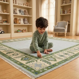 May include: A large square area rug featuring a light blue centre field surrounded by a wide cream-coloured border decorated with a repeating green and blue floral and leaf pattern, finished with short white fringe along the edges.