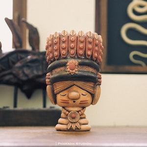 Puede incluir: Figura de cerámica de una figura nativa americana estilizada con los ojos cerrados. La figura es de color canela con un tocado y detalles en verde oscuro y marrón. El tocado presenta detalles en forma de plumas. La figura tiene un collar y es probablemente un objeto decorativo.