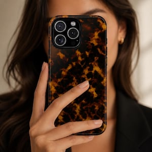 Op de afbeelding: Een telefoonhoesje met een schildpadpatroon, voor het gezicht van een persoon gehouden. De hoes heeft een donkerbruin en amberkleurig ontwerp. De cameralenzen van de telefoon zijn zichtbaar. De persoon draagt een zwarte blazer.