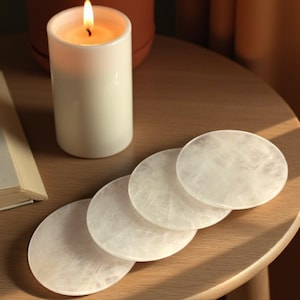 May include: A lit white candle and four round, white, translucent coasters are arranged on a small, round, wooden table. A potted plant and an open book are also on the table. The scene is lit by natural light.