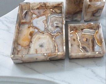 Op de afbeelding: Een badkamerset met een zeepdispenser, tandenborstelhouder en twee trays. De set is gemaakt van agaatsteen in beige, bruine en witte tinten, met gouden accenten. De items zijn geplaatst op een marmeren aanrecht.