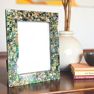 Könnte beinhalten: Ein dekorativer Bilderrahmen mit einem Mosaik-Stil-Rand in Grün-, Blau- und Goldtönen. Der Rahmen steht auf einer dunklen Holzoberfläche, mit einem weißen Innenraum. Eine Vase mit Zweigen und Büchern befindet sich im Hintergrund.