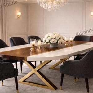 May include: An oval dining table with a white and wood grain top, supported by a gold-coloured X-shaped base. Eight grey upholstered chairs with dark wood legs and gold accents surround the table. A chandelier hangs above, illuminating the dining area.