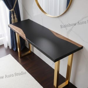Può includere: Una consolle moderna con piano nero e dettagli in legno naturale. Il tavolo ha gambe in metallo color oro. È posizionata davanti a una parete bianca con uno specchio dorato e tende blu.