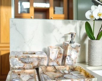 Op de afbeelding: Badkameraccessoireset met een tissuebox, zeepdispenser, beker, dienblad en zeepbakje. De set is gemaakt van een steenachtig materiaal met grijze, beige en witte kleuren. Een witte vaas met orchideeën en een chromen kraan zijn ook zichtbaar.