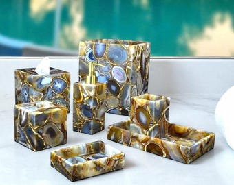 Op de afbeelding: Een badkamerset van agaatsteen, met een zeepbakje, tissuebox en lotiondispenser. De steen heeft blauwe, bruine en gouden tinten. Een opgerolde witte handdoek en een glazen diffuser met zwarte stokjes zijn ook aanwezig.