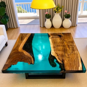 Może przedstawiać: Kwadratowy stolik kawowy o unikalnym designie. Stół ma drewniany blat z turkusowym środkiem z żywicy. Drewno ma naturalny wzór słojów. Jest wspierany przez czarną metalową ramę.