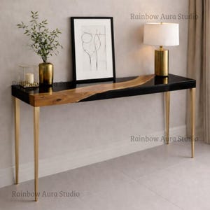 Puede incluir: Una consola moderna con una tapa negra brillante y una incrustación de madera natural. La mesa tiene patas de metal dorado y está decorada con una lámpara, obras de arte y un jarrón. La mesa mide aproximadamente 122 cm de largo.