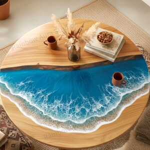 Puede incluir: Mesa de centro redonda de madera con diseño de océano de resina azul. La mesa presenta una tapa de grano de madera natural con un vibrante patrón de olas azules y blancas. Se ven libros, un cuenco de nueces y tazas marrones.