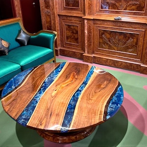 May include: A round coffee table with a unique design. The table features a wooden surface with blue resin accents, creating a river-like effect. The table is supported by a pedestal base and is placed in a room with a teal sofa.
