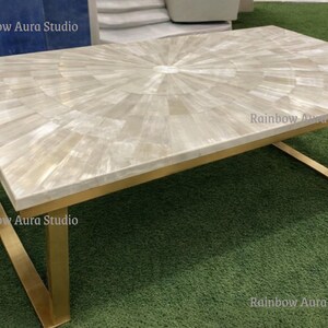 May include: A rectangular coffee table with a light-colored, patterned top and gold-colored metal legs. The tabletop features a radial design. The legs are angled and create a modern aesthetic. The table is set on a green surface.