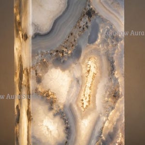 May include: A rectangular, illuminated lamp made of a translucent, patterned stone. The stone features swirling patterns in shades of white, gray, and brown, with a warm, glowing light emanating from within. The lamp sits on a dark, rectangular base.
