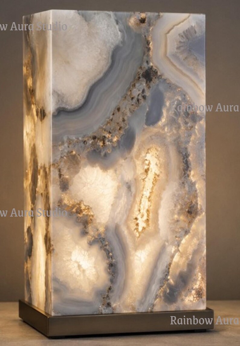 May include: A rectangular, illuminated lamp made of a translucent, patterned stone. The stone features swirling patterns in shades of white, gray, and brown, with a warm, glowing light emanating from within. The lamp sits on a dark, rectangular base.
