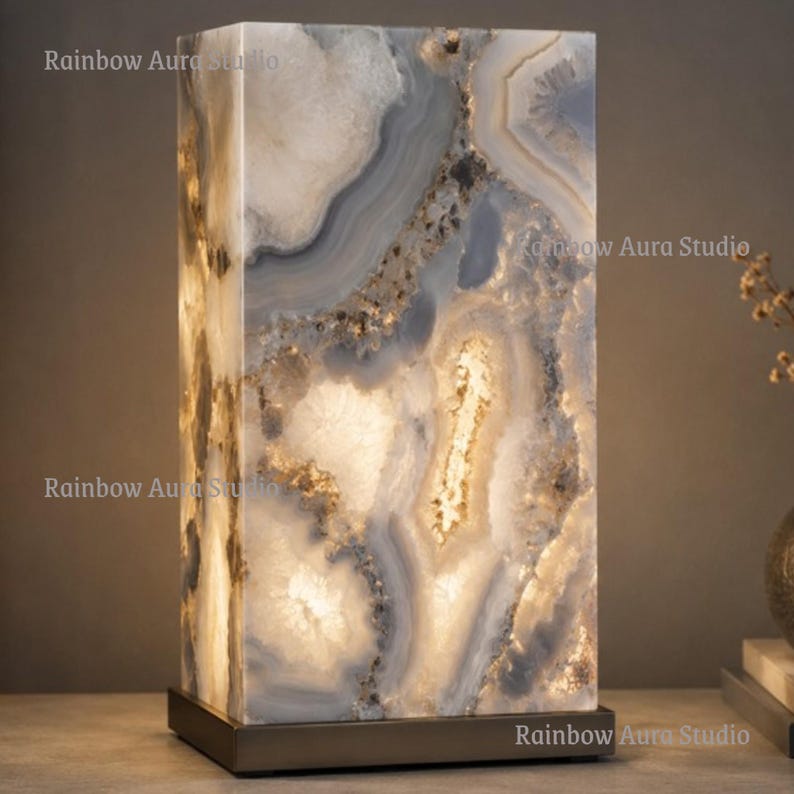 May include: A rectangular table lamp with a natural stone design. The lamp features a base and a shade with a pattern of white, gray, and brown swirls. The lamp is lit from within, creating a warm glow. The lamp is a decorative home decor item.