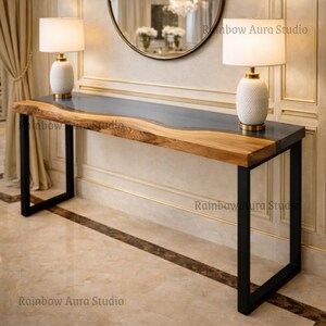 May include: A long, rectangular console table with a live edge wood top and a dark gray resin inlay. The table has black metal legs and two white ceramic lamps with gold accents. The text "Rainbow Aura Studio" is visible.