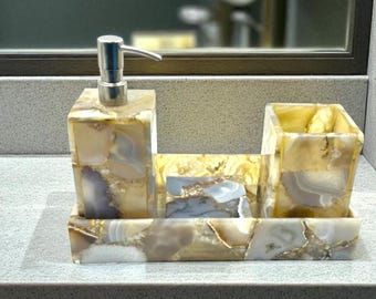 Op de afbeelding: Een badkameraccessoireset gemaakt van agaatsteen. De set bevat een zeepdispenser met een zilveren pomp, een zeepbakje en een beker, allemaal op een bijpassend dienblad. De steen heeft een mix van beige, wit en paars.