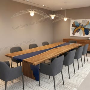 May include: A large wooden dining table with a blue resin river design, surrounded by gray upholstered chairs. The table is in a modern dining room setting with a large abstract painting and decorative lighting.
