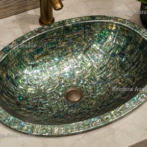 May include: Oval-shaped bathroom sink with a mosaic-like pattern in shades of green, blue, and iridescent hues. The sink has a metallic drain and is set on a light-colored countertop. The text "Rainbow Aura Studio" is visible.