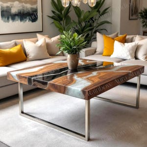 Op de afbeelding: Een rechthoekige salontafel met een blad van hout en hars, met een vloeiend patroon van bruin, zwart en wit. De tafel heeft een gepolijste afwerking en staat op een frame van geborsteld metaal. De kamer omvat een grijze bank met gele en witte kussens.