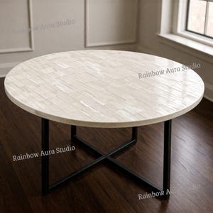 Op de afbeelding: Een ronde salontafel met een wit, mozaïekachtig blad en zwarte metalen poten. Het tafelblad is gemaakt van rechthoekige stukken, waardoor een textuur ontstaat. De poten vormen een X-vorm aan de basis. De tafel staat op een donkere houten vloer.