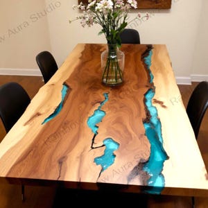 Puede incluir: Mesa de comedor rectangular de madera con incrustaciones de resina turquesa en forma de río. La mesa está rodeada de sillas negras y coronada por un jarrón con flores. Dimensiones: aproximadamente 183 cm de largo y 91 cm de ancho.