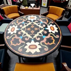 May include: A round table with a black surface, adorned with a floral mosaic pattern in white, orange, and blue. The table is surrounded by armchairs with orange and navy blue upholstery, and red accent pillows.