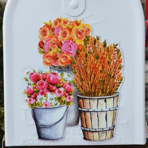 May include: White mailbox with a floral design. The design features three buckets with different types of flowers. The flowers are painted in shades of pink, orange, and yellow.