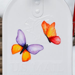 May include: White metal mailbox featuring a decorative design of two butterflies. The butterflies are painted in shades of orange, yellow, purple, and red. The mailbox has the text "US MAIL" embossed on the front.
