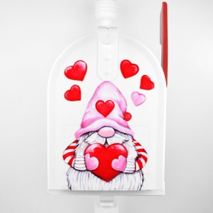 May include: White mailbox with a red flag, decorated with a gnome holding a red heart. The gnome is wearing a pink hat with a red heart on it. There are several red hearts scattered around the gnome.