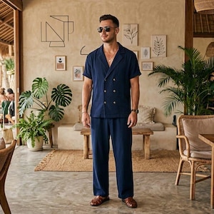 May include: A navy blue short-sleeved suit with gold buttons. The double-breasted shirt and matching pants are worn with brown leather sandals. The outfit is worn in a cafe setting with plants and wooden furniture.