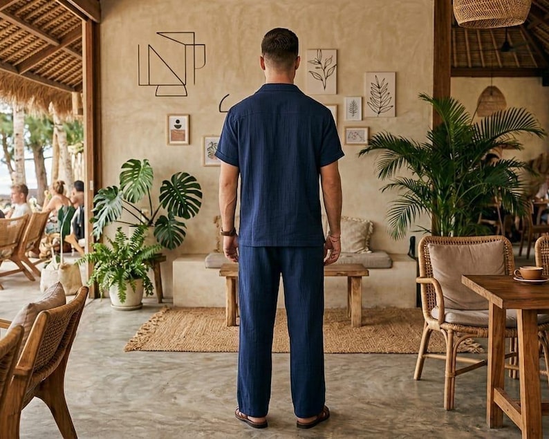 May include: A person wearing a navy blue short-sleeved shirt and matching pants. The outfit is a casual style, suitable for warm weather. The person is standing in a cafe or restaurant setting, with natural light and decor.